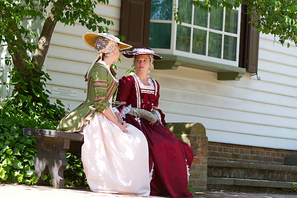Two women in historical dresses sit on a bench, chatting in a sunny garden. One wears green and pink, the other red. Wooden house background.