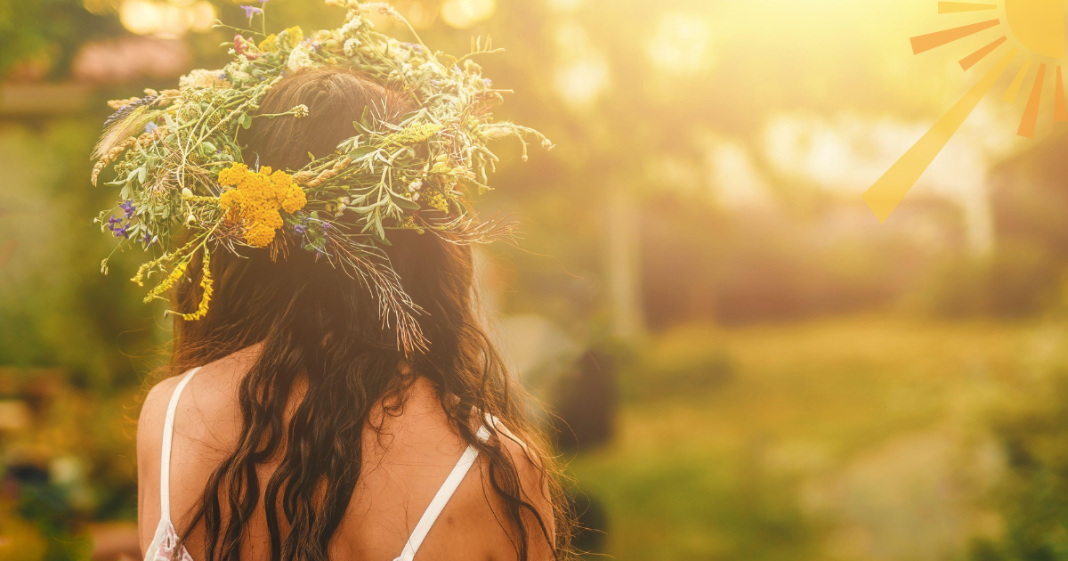 A woman with long brown hair wearing a flower crown facing a sun drenched meadow